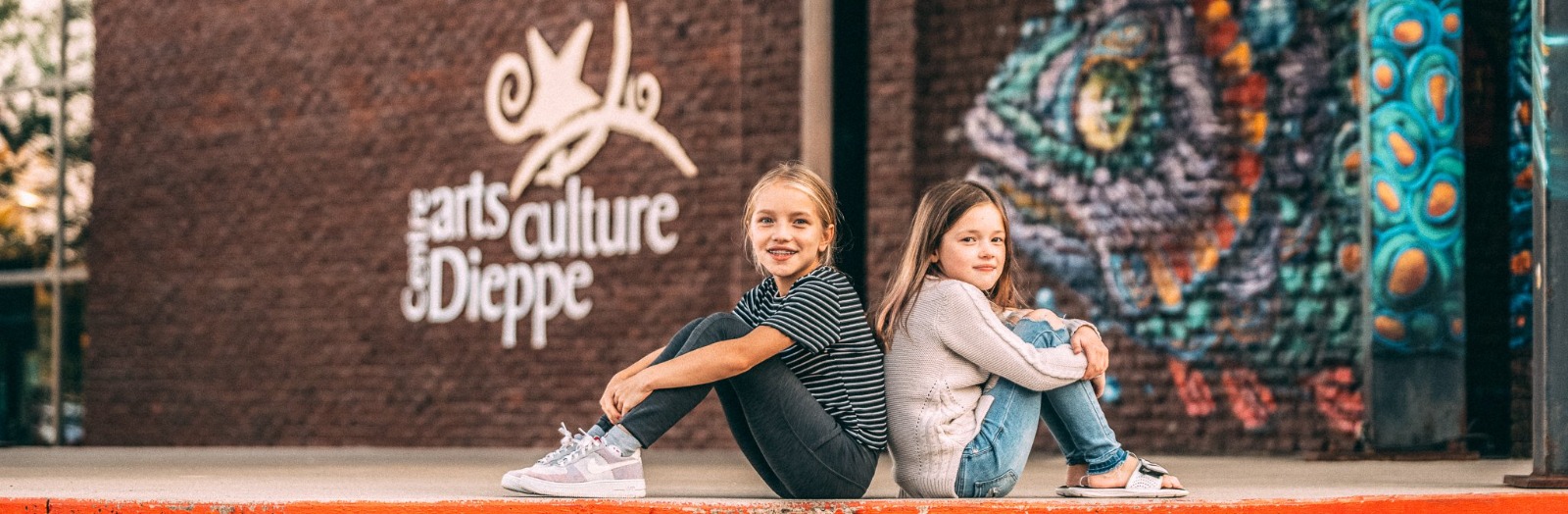 Two girls sitting in front of the Dieppe Arts and Culture Centre's sign